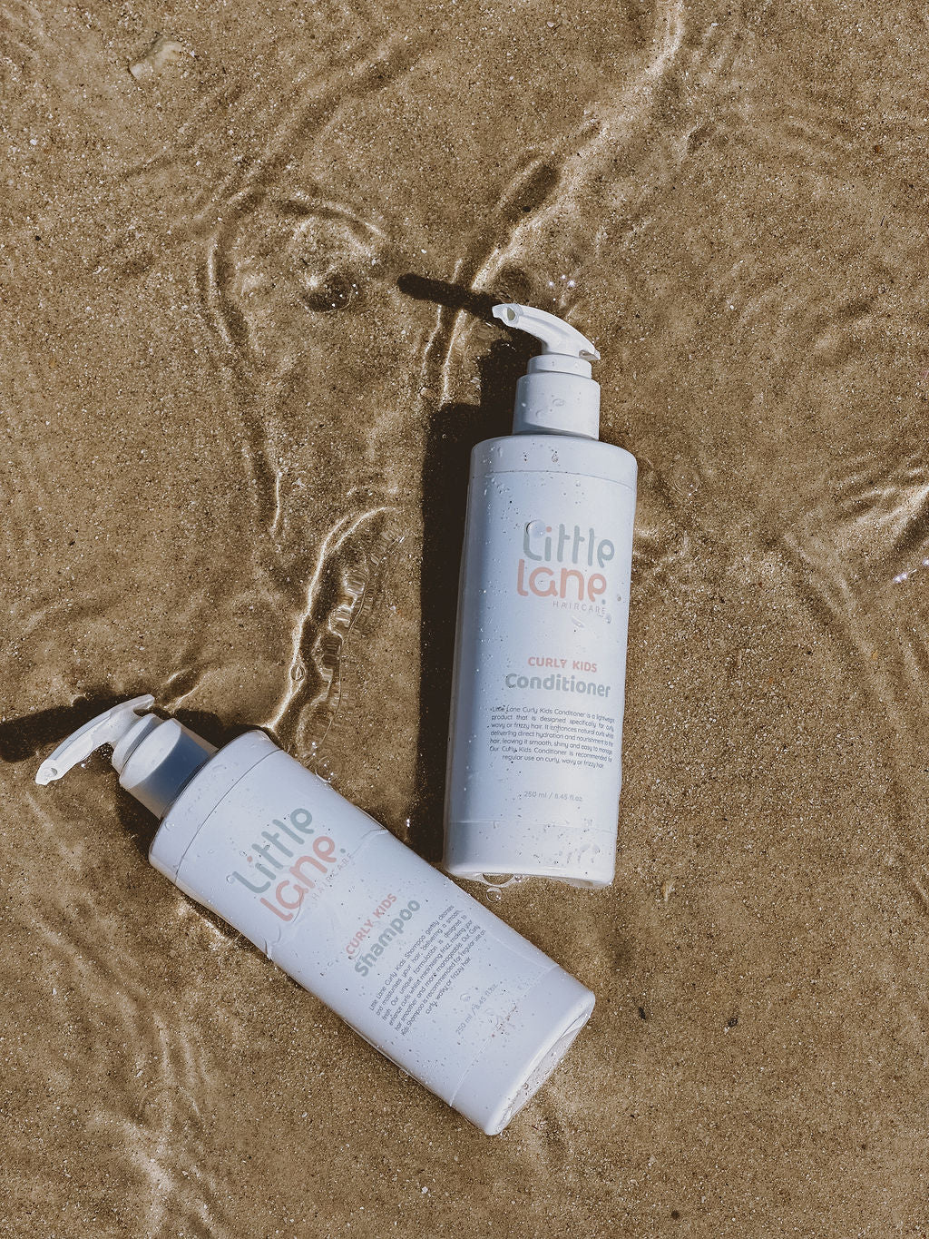 Two bottles of 'Little Lane' shampoo and conditioner on wet sand.