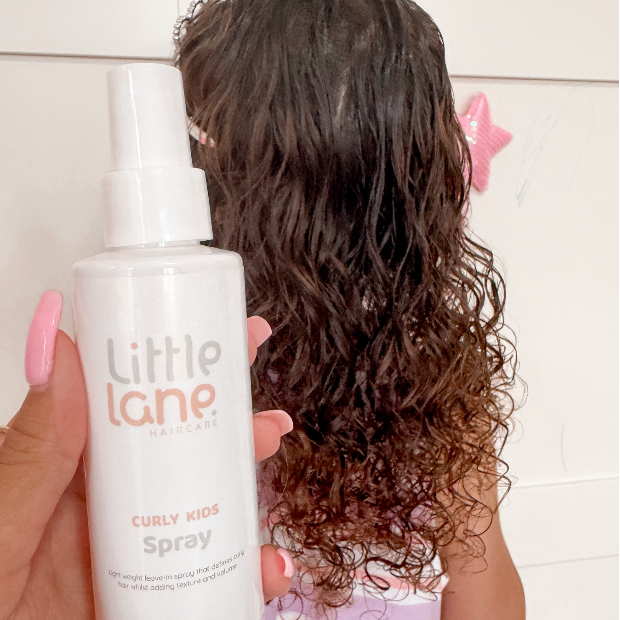 Person holding a 'Little Lane' curly kids spray bottle with a child's wavy hair in the background.